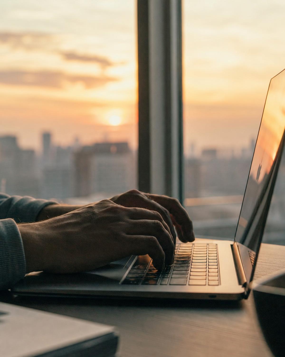 Hands typing on a laptop with warm sunset light through a city window